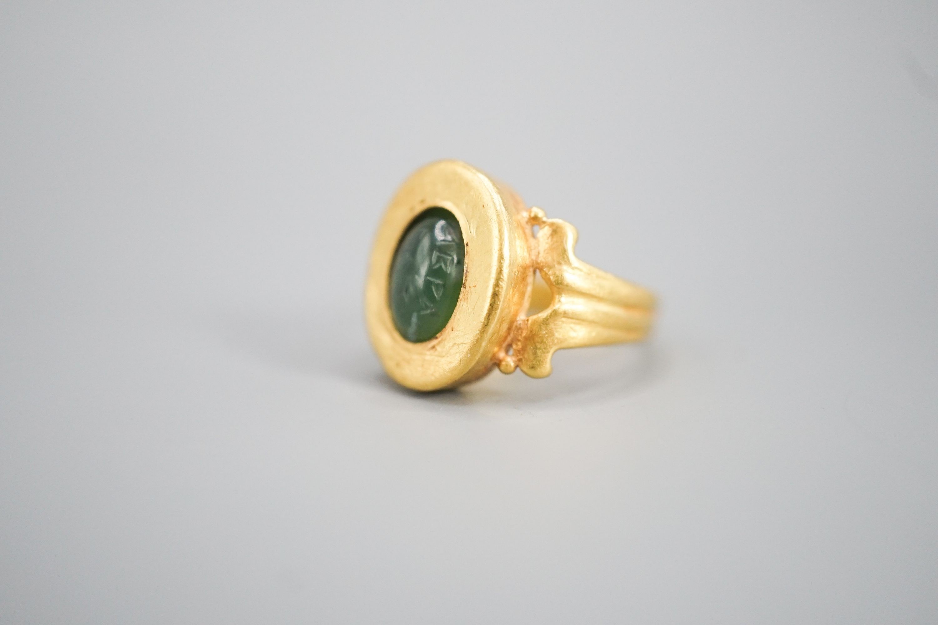 An antique yellow metal and carved nephrite? oval ring, size H, gross 4 grams (the stone is loose in situ and needs re-setting).